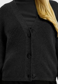 Cardigan en tricot noir avec un col en V, présentant un motif texturé, trois boutons à l'avant et des poignets et un ourlet côtelés.