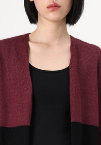 Cárdigan de punto burdeos con abertura frontal, combinado con una blusa negra ajustada. Tejido texturizado con un acabado suave y dobladillo negro en contraste.
