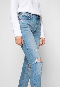 Vaqueros rotos de color azul claro, hechos de mezclilla, con una cintura alta y detalles desgastados en las rodillas. Combinados con una sudadera blanca.