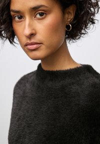 Schwarzer, flauschiger Strickpullover mit hohem, rundem Halsausschnitt. Die Textur ist weich und hat einen dezenten Glanz, der durch einfache goldene Creolen ergänzt wird.