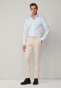 Camicia button-down azzurra con maniche lunghe, abbinata a pantaloni color crema e scarpe scure in suede, in piedi davanti a uno sfondo neutro.
