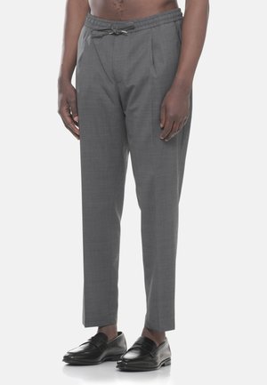 Uomo che indossa pantaloni grigi su misura con coulisse e mocassini in pelle neri, in piedi davanti a uno sfondo chiaro e semplice.