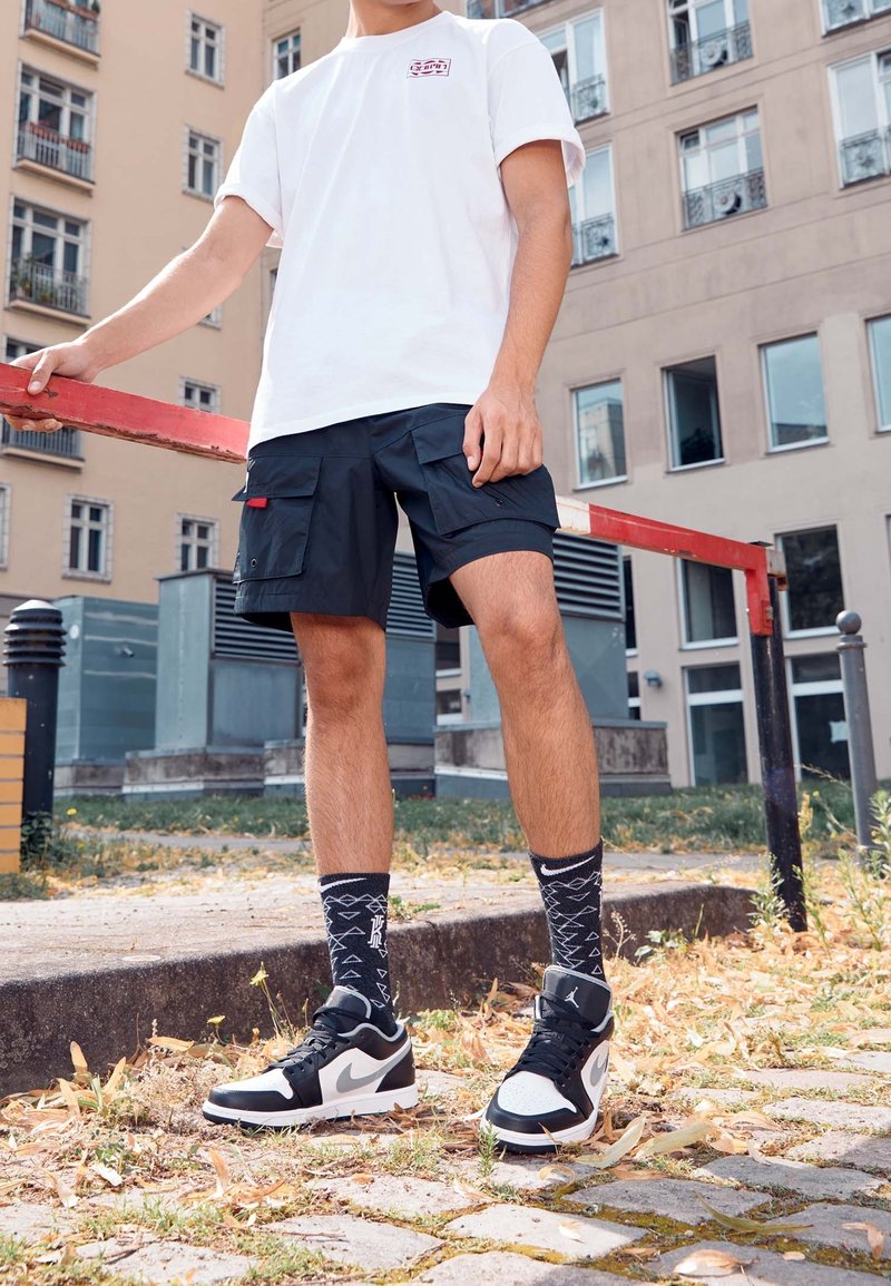 Bermudas negras con múltiples bolsillos, camiseta blanca, calcetines negros con patrones y zapatillas negras y blancas en un entorno al aire libre.