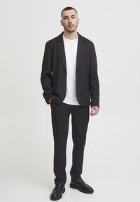Black tailored blazer with a single chest pocket, paired with black trousers and a white t-shirt. Man wearing black dress shoes stands upright.