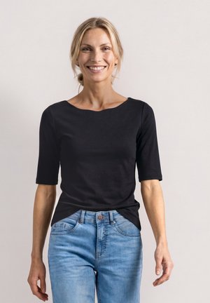 Lächelnde Frau mit blond zurückgebundenem Haar, die ein schwarzes Halbarmshirt und hellblaue Jeans trägt, vor einem schlichten hellen Hintergrund stehend.
