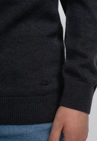 Donkergrijze trui met een geribde zoom en manchetten. Bevat een klein zwart merkteken. Zachte textuur, aansluitend ontwerp, gedeeltelijk zicht op de mouw en hand.