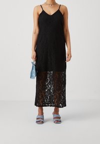 Black lace maxi dress featuring a fitted top with spaghetti straps, a semi-sheer lower section, and a v-neckline. Accompanied by blue accessories.