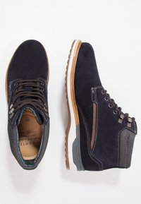 Navy Wildleder-Ankle Boots mit braunen Schnürsenkeln, hellbraunen Akzenten und einer weißen Mittelsohle. Strukturiertes Seitenpaneel und Logo auf der Sohle für zusätzliche Details.