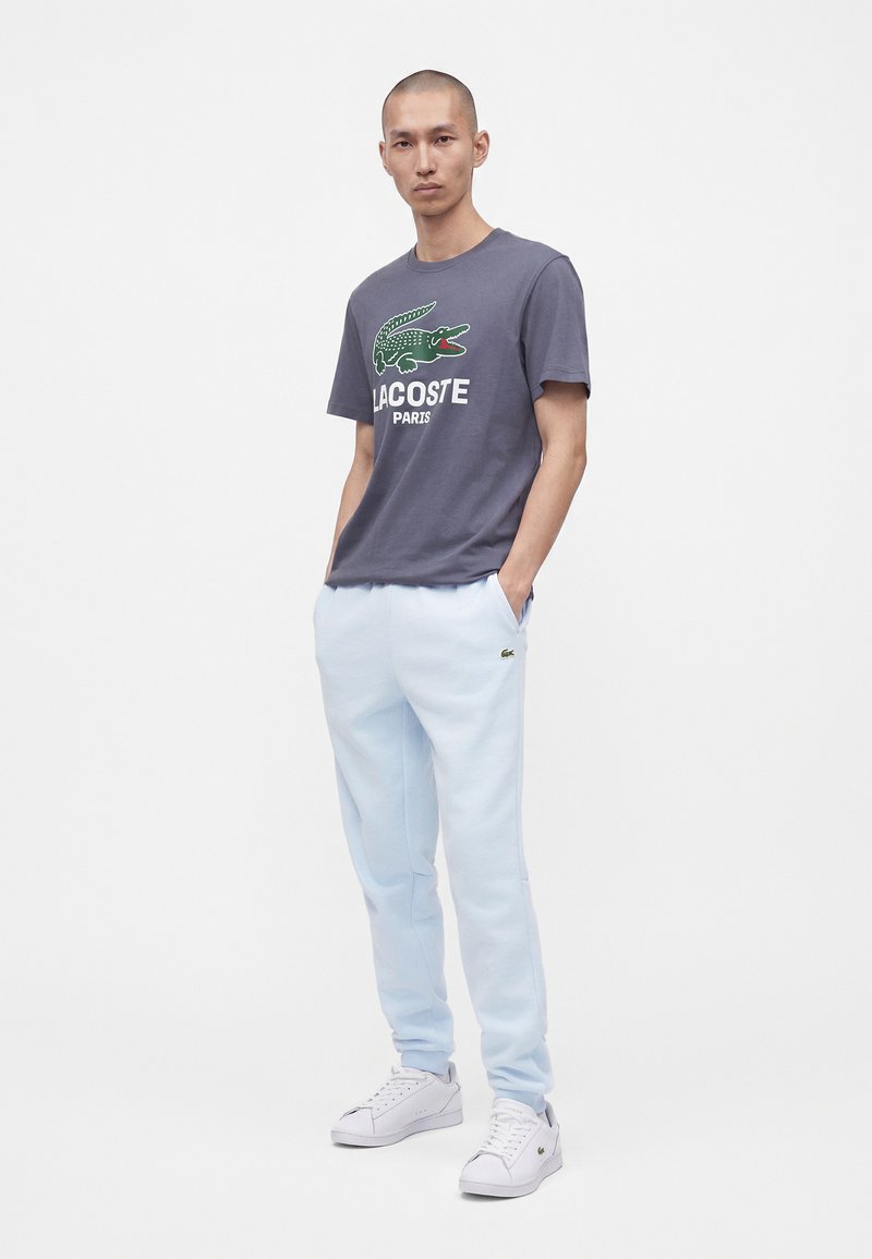 Άνδρας με γκρι μπλουζάκι Lacoste με πράσινο λογότυπο κροκοδείλου, ανοιχτό μπλε φόρμες και λευκά αθλητικά παπούτσια, στέκεται με τα χέρια στις τσέπες.