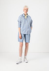 Blue and white striped button-up shirt, rolled sleeves, paired with light denim shorts. Model wears white sneakers and ankle socks.