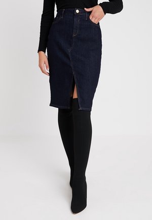 Vrouw draagt een donker denim knielange rok met een voorin gemaakte split, gecombineerd met zwarte panty's en een zwart top met lange mouwen, hand in de zak van de rok.