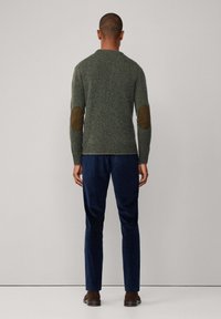 Ein Mann steht mit dem Rücken zur Kamera, trägt einen grünen Pullover mit braunen Ellbogenflicken, marineblaue Hosen und braune Schuhe vor einem schlichten Hintergrund.