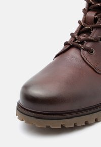 Hnědá kožená bota s kulatou špičkou, texturovaným povrchem, kontrastními hnědými tkaničkami a robustními, béžovými gumovými podrážkami s výrazným vzorem. 