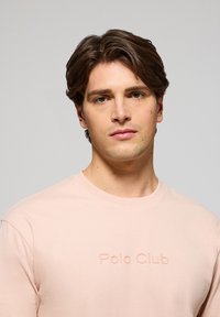 Camiseta de algodón de color rosa claro con un logo en relieve de "Polo Club" en el pecho. Cuello redondo y mangas cortas. Textura suave.