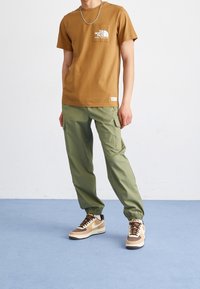 Brązowy bawełniany t-shirt z logo na przodzie, zielone spodnie cargo z elastycznymi mankietami oraz wielokolorowe sneakersy w odcieniach białym, brązowym i beżowym.