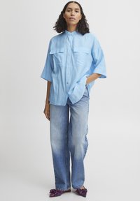 Camicia azzurra chiara con maniche corte e due tasche sul petto, abbinata a jeans in denim sbiaditi e larghi e ballerine a fantasia.