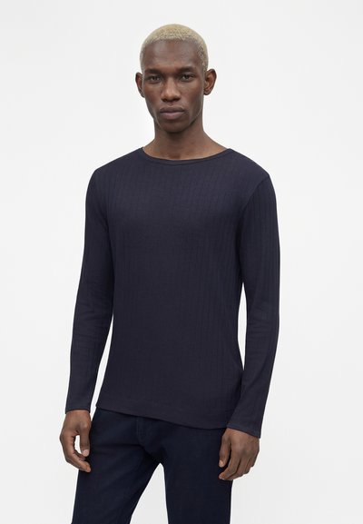 Νέος άνδρας με κοντά ξανθά μαλλιά, φοράει μπλε σκούρο ριμπ μακρυμάνικο πουκάμισο και σκούρο παντελόνι, στέκεται μπροστά σε λευκό φόντο.