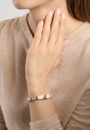 Goldfarbener Armreif mit abwechselnd rechteckigen Gliedern und einem runden Emblem, getragen an einer Hand vor einem sanften, beigen Pulloverhintergrund.