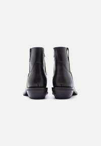 Μαύρα δερμάτινα μποτάκια αστραγάλου, εμφανιζόμενα από πίσω, με χαμηλά τακούνια και θηλιές για τράβηγμα σε λευκό φόντο.