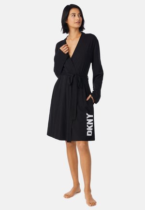 Vrouw draagt een zwarte knielange badjas, gestrikt in de taille met witte DKNY letters aan de onderzijde, staat blootsvoets op een effen achtergrond.