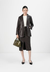 Vrouw in een donkerbruine leren blazer en rok, met een wit getextureerd topje, zwarte hakken, die een olijfgroene handtas vasthoudt tegen een witte achtergrond.