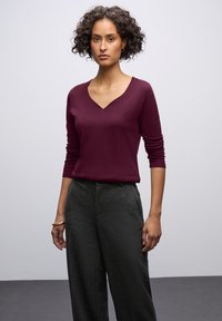 Burgunderfarbener V-Ausschnitt-Pullover mit Dreiviertelärmeln, kombiniert mit taillierten dunkelgrauen Hosen mit subtiler Textur.