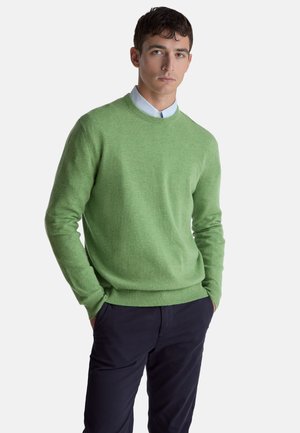 Zelený svetr z měkké látky, s klasickým kulatým výstřihem a žebrovanými manžetami. Kombinován s tmavými kalhotami, na bílém pozadí.