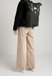 Ciemnozielony plecak z płótna z zapięciem na klapę, regulowanymi paskami, srebrnym okuciem i przednią metką. Nosi go osoba, zestawiony z beżowymi spodniami.