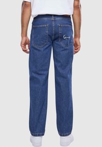 Mørkeblå denimjeans med rett ben-design, to baklommer og hvit brodert logo på høyre lomme. Slik tekstur.