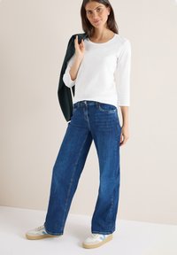 Witte long-sleeve top met een ronde halslijn, gecombineerd met loszittende donkerblauwe jeans en beige sneakers, met een groene jas over de schouder geslagen.
