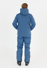 Blå isolerad skidjacka med huva och matchande byxor. Har en mjuk textur, skräddarsydd passform och kontrasterande svarta accenter vid byxbenens fåll.