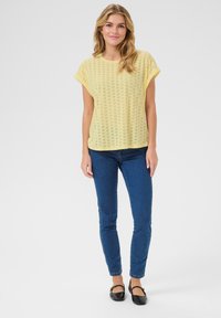 Gelbes Kurzarmoberteil mit Lochmuster, kombiniert mit blauen engen Jeans und schwarzen Ballerinas. Das Design ist leicht und lässig.