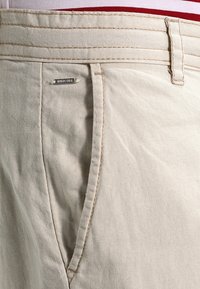 Pantalones de lino beige con bolsillo lateral, costura visible, trabilla para cinturón y pequeña etiqueta metálica etiquetada "INDICODE".