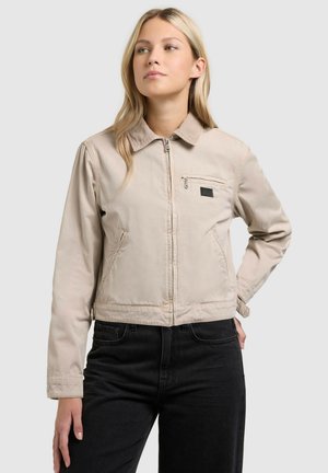 Naine, kes kannab beeži värvi lukuga jope krae ja rinnataskuga, mida ta kannab koos mustade teksapükstega, seisab ükskõikse helehalli tausta taustal.