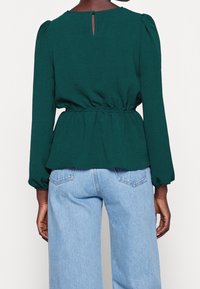 Blouse texturée bleu sarcelle à coupe décontractée, manches bouffantes et ouverture en forme de goutte d'eau dans le dos, associée à un jean large bleu clair.