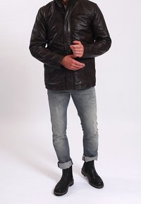 Braune Lederjacke mit Stehkragen, frontalem Reißverschluss und zwei seitlichen Taschen. Kombiniert mit hellblauen, verwaschenen Jeans und schwarzen Stiefeletten.