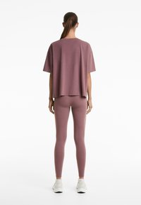 Femme de dos, portant un T-shirt oversize mauve et un legging assorti, avec des baskets blanches, sur un fond blanc uni.