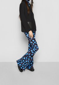 Blazer negro sobre pantalones acampanados de patrón floral azul que presentan flores azules y rosas sobre un fondo oscuro. Tela suave, silueta ajustada.