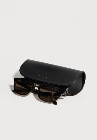 Γυαλιά ηλίου με σχέδιο καβούκι χελώνας και σκούρους φακούς, τοποθετημένα μπροστά από μια μαύρη σκληρή θήκη από δέρμα σε λευκή επιφάνεια.