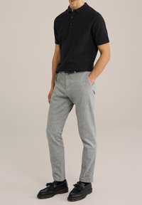 Czarna koszulka polo z krótkim rękawem wykonana z teksturowanej tkaniny, zestawiona z jasnoszarymi spodniami. Model nosi czarne buty z ozdobnymi frędzlami.