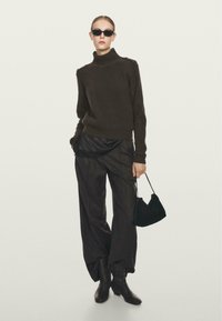 Maglione a collo alto a maglia marrone con dettagli sulle spalle, pantaloni neri larghi, borsa nera e stivali neri alti, modella che indossa occhiali da sole.