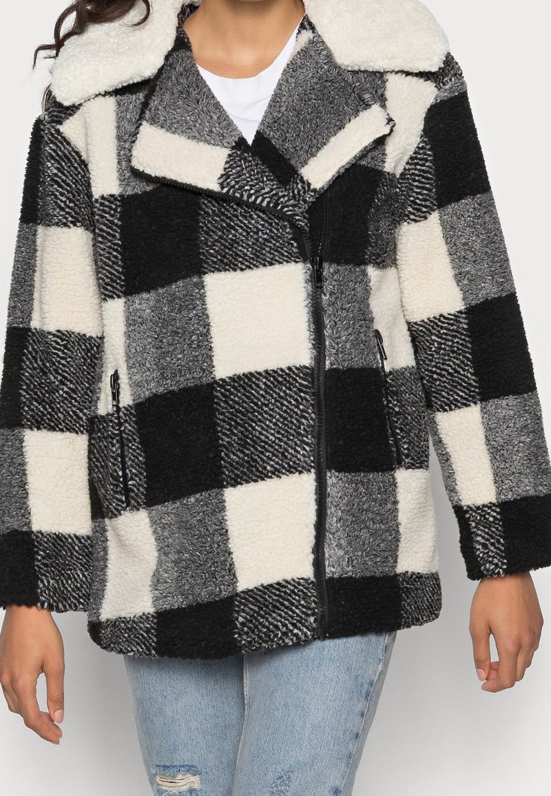 Frau trägt eine schwarze, weiße und graue karierten Fleecejacke mit breitem Kragen und mit Reißverschlüssen versehenen Taschen, kombiniert mit hellblauen, zerrissenen Jeans.