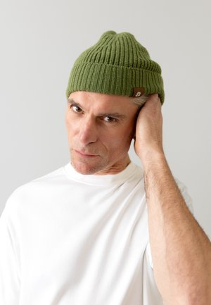 Groene gebreide beanie met verticale ribbels en een klein bruin label. Model draagt een wit t-shirt en heeft een neutrale uitdrukking.