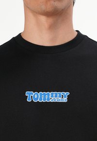 Man in een zwart T-shirt met ronde hals, met het "Tommy Jeans" logo in het blauw en wit gecentreerd op de borst.