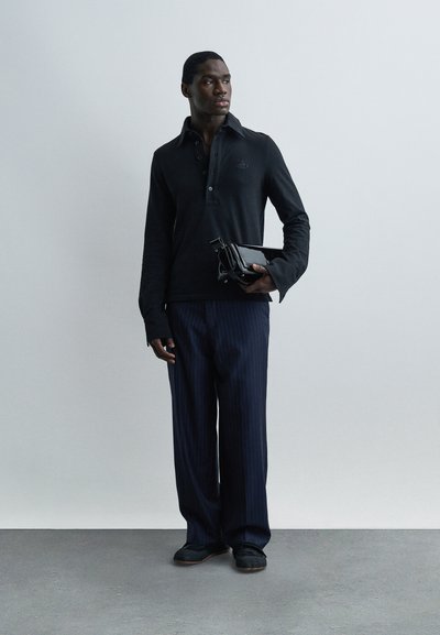 Άνδρας που στέκεται μπροστά σε ένα απλό τοίχο, φοράει μαύρο μακρυμάνικο πόλο, μπλε παντελόνι με ρίγες, μαύρα παπούτσια και κρατά μια μαύρη τσάντα χειρός.