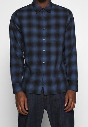 Man draagt een blauw-zwart geruit overhemd met lange mouwen en donkere jeans, staand tegen een eenvoudige achtergrond.