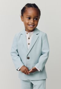 Lichtblauw pak met een enkele knoop, twee voorzakken en een gladde textuur. Wit ondershirt met knoopdetails. Kind staat lachend.