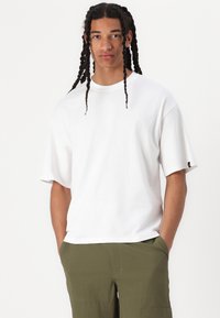 T-shirt en coton blanc avec une coupe décontractée, des manches courtes, un col rond et un petit logo sur la manche. Associé à un pantalon vert olive.