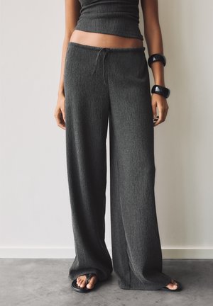 Femme portant un pantalon large texturé gris foncé, un haut sans manches assorti, des bracelets noirs et des sandales noires, debout à l'intérieur.