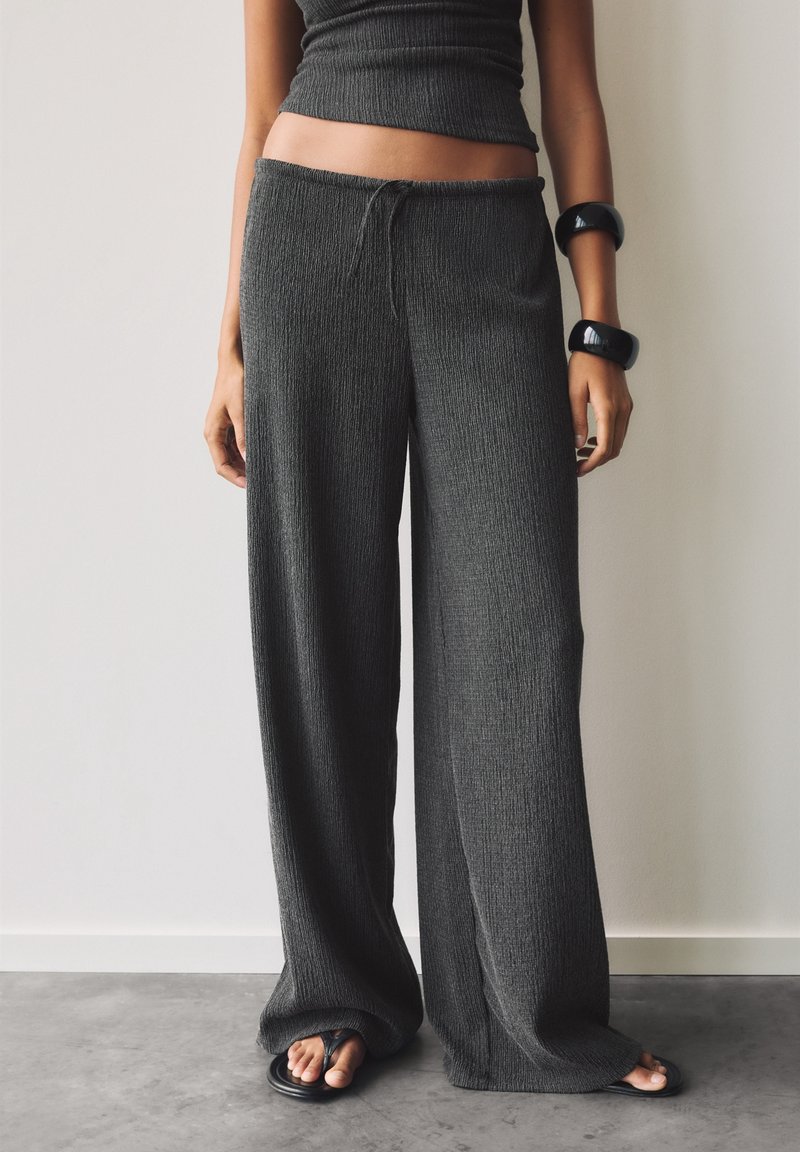 Femme portant un pantalon large texturé gris foncé, un haut sans manches assorti, des bracelets noirs et des sandales noires, debout à l'intérieur.